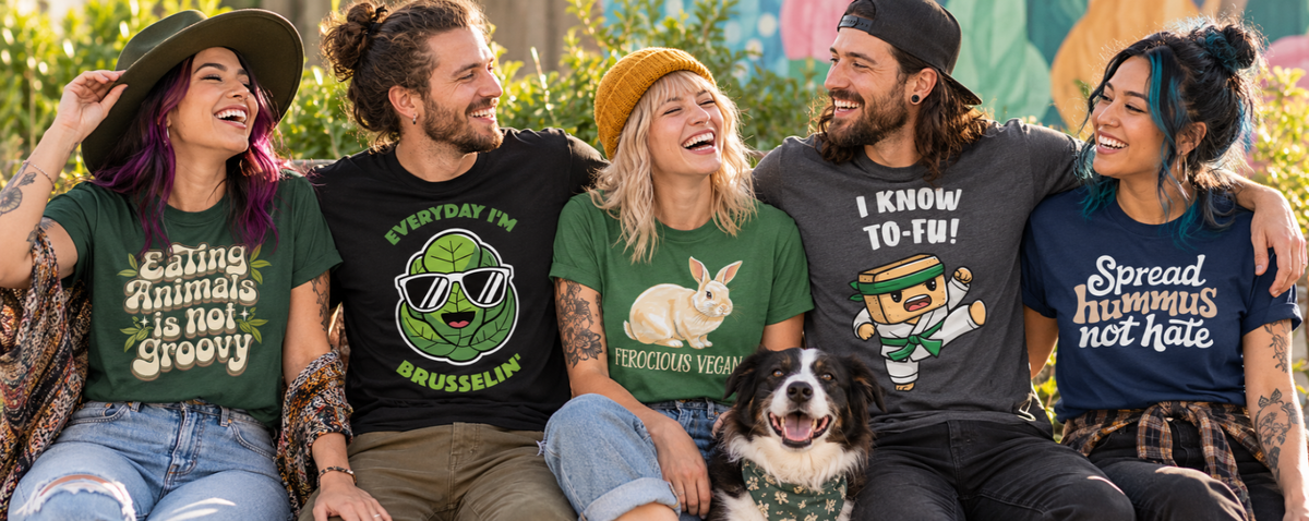 Five people sitting together outdoors wearing graphic t-shirts with humorous and playful designs, accompanied by a black and white dog. The shirts feature various green, black, and blue colors with text like 'Eating animals is not Groovy', 'Everyday I'm BRUSSELING', 'Ferocious Vegan' with a bunny graphic, 'I KNOW TO-FU!', and 'Spread hummus not hate',